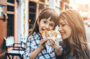 Mom with her 6 years old daughter walking along city street and eating ice cream in front of the outdoor cafe.