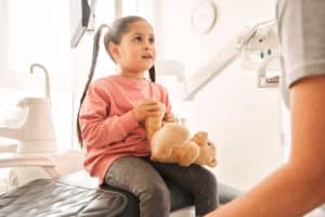 smiling little girl sitting in dental chair listening to female dentist holding teddy bear